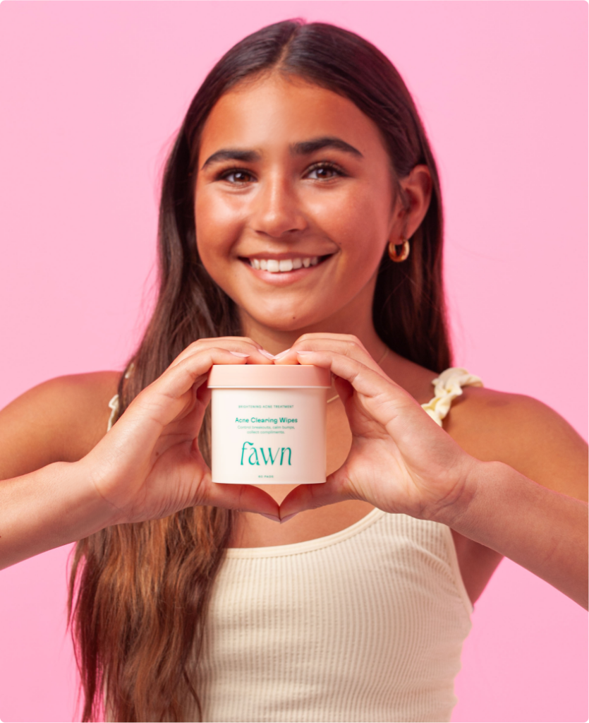 A teen girl smiling and holding Fawn Acne Wipes on a pink background.